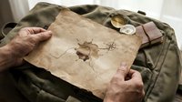 Mature male hands holding aged parchment map with compass and route markings over military canvas fabric, suggesting adventure planning and exploration themes.