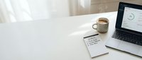 Clean white desk workspace with laptop displaying analytics dashboard, coffee cup and notepad for productive morning routine and business planning.