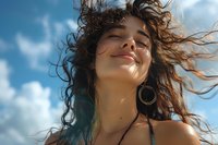 Young Hispanic woman with curly hair blowing in wind, eyes closed with serene expression, wearing hoop earrings against blue sky with white clouds, natural sunlight.