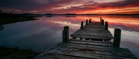 Wooden pier extending into calm lake at sunset with vibrant orange and purple sky reflected in still water, serene landscape for travel and nature projects.