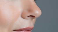Close-up profile view of Caucasian female nose and lips showing natural skin texture and pores against neutral gray background, suitable for beauty and skincare marketing.