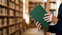 Person holding green hardcover book in library with shelves of books blurred in background. Educational and reading concept for academic, publishing, and learning materials.