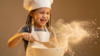Young girl chef with braids wearing white hat and apron mixing ingredients in bowl with flour dust flying, joyful cooking experience in warm kitchen.