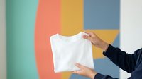 Hands holding blank white t-shirt against colorful geometric background with vertical stripes in mint, coral, yellow and blue tones for apparel mockup and branding.