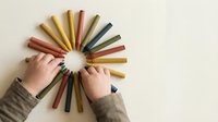 Colorful wooden crayons arranged in circle with child hands reaching toward center on neutral background for creative art education projects.