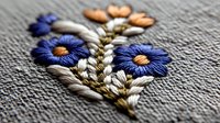 Delicate embroidered floral pattern featuring blue cornflowers and peach blooms on gray linen fabric, showcasing detailed needlework and traditional stitching techniques.