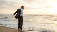 Professional businessman standing on beach at sunset holding jacket over shoulder looking at ocean waves during golden hour vacation or work-life balance moment.
