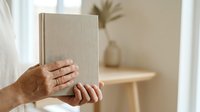 Mature adult with medium skin tone holding closed beige linen book against neutral interior background with natural window light and blurred home decor.