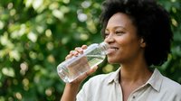 Happy African American woman drinking fresh water from glass bottle outdoors in natural green park setting for healthy lifestyle and wellness.
