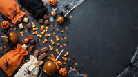 Halloween candy and treats scattered on dark slate background with orange gift bags, plastic spiders, pumpkins and festive decorations for autumn celebration.