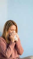 Woman with shoulder-length brown hair, Caucasian ethnicity, wearing mauve sweater, blowing nose into tissue against blue background. Healthcare, illness, cold, allergy concept.