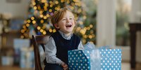 Joyful young boy laughing with excitement while holding blue polka dot gift box during Christmas celebration with decorated tree in background.