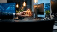 Businessman hand touching glowing warning triangle hologram at modern office desk with multiple monitors displaying data analytics and charts.