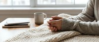 Cozy winter morning at home, woman in knit sweater with clasped hands resting on blanket near coffee mug and notebook on wooden table.