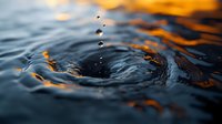 Water drops falling into dark rippling surface creating concentric circles with golden sunset reflections dancing on waves. Artistic macro photography for nature themes.