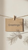 Kraft paper card with Greetings text hanging on twine with clothespin, pine branch shadow on beige wall, minimal holiday concept.