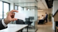 Businessman holding blank white business card in modern office environment with colleagues working in background.