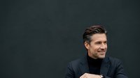 Middle-aged Caucasian male professional wearing dark navy jacket and black turtleneck, smiling confidently against dark background. Corporate portrait for business, leadership, and professional use.