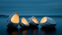 Illuminated stone sculptures with glowing warm light emerging from carved openings, arranged in row against moody blue twilight seascape background.