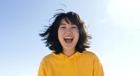 Young woman with dark shoulder-length hair, East Asian ethnicity, wearing bright yellow hoodie, smiling joyfully outdoors against clear sky background.