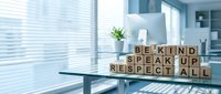 Wooden letter blocks spelling BE KIND SPEAK UP RESPECT ALL on modern office desk near window with natural light and plants in professional workspace.