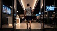 Young man walking through modern gym corridor with equipment and screens visible in fitness center interior.