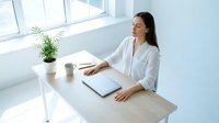 Young professional woman working on laptop at modern white desk by window with plant and coffee cup in bright minimalist office space for remote work.