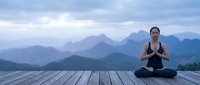 Young Asian woman practicing yoga meditation in lotus pose on wooden deck with misty mountain landscape background at dawn for wellness and mindfulness concepts.