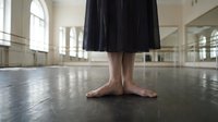 Barefoot ballet dancer standing in a studio, black skirt, wooden floor, barre and mirrors in background, graceful dance training moment.