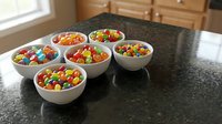 Six white bowls filled with colorful candy-coated chocolates arranged on dark granite kitchen counter for party snacks and sweet treats.