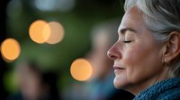 Serene mature woman with silver hair in profile, eyes closed peacefully against soft bokeh lights background, wearing blue knit sweater, expressing tranquility and mindfulness.