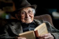 Elderly Caucasian man in hat and glasses reading book while relaxing in armchair, wearing warm knit sweater and tweed jacket, expressing joy and contentment.