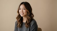 Young Asian woman with long wavy brown hair smiling warmly in casual gray sweater against neutral beige background for lifestyle and portrait photography.