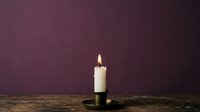 Lit white candle in dark holder on wooden surface against deep purple background, suitable for wellness, spirituality, and memorial designs.