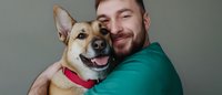 Heartwarming moment between smiling mixed breed dog wearing red collar and bearded man in green shirt embracing, showing bond between pet and owner against gray background.