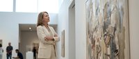Professional woman in beige blazer contemplating abstract artwork in modern gallery space with natural lighting and contemporary interior design.