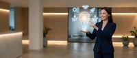 Professional Asian businesswoman in dark suit presenting innovative ideas with digital holographic interface displaying lightbulb and icons in modern office lobby.