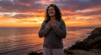 Young Hispanic woman with curly hair practicing mindfulness meditation at ocean sunset, hands on heart in gratitude pose against dramatic golden sky and waves.