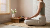 Young woman practicing meditation in serene home setting with wooden tea table, flowers and natural light streaming through bamboo blinds.