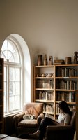 Cozy reading nook with young Asian woman enjoying book in leather armchair beside arched window and wooden bookshelf filled with pottery and literature.