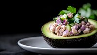 Fresh diced tuna with red onion and parsley served in halved avocado on white plate against dark background, healthy appetizer with raw fish.