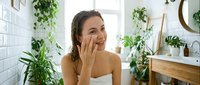 Young woman applying skincare cream in modern bathroom with plants, natural beauty routine and self-care wellness lifestyle.