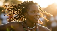 Young African woman with flying dreadlocks and colorful beaded necklaces dancing outdoors in golden sunset light, joyful expression.