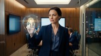 Young woman with dark hair and fair complexion, wearing professional dark blazer, interacting with holographic facial recognition technology in modern corporate office environment with wood paneling and digital displays.