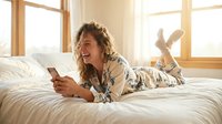 Young woman with curly blonde hair and fair complexion lying on bed in sunlit bedroom, wearing casual patterned shirt, holding smartphone, smiling while relaxing at home.