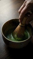 Male hand whisking matcha powder in ceramic bowl with bamboo whisk, traditional Japanese tea ceremony preparation on dark surface.