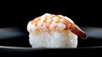Fresh shrimp nigiri sushi piece on black plate with soft lighting, showcasing traditional Japanese cuisine preparation and professional food photography.