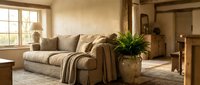 Cozy living room with beige upholstered sofa, warm natural lighting from large windows, potted plant, and neutral earth-tone decor for home interior design and lifestyle photography.
