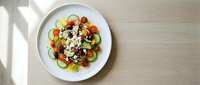 Fresh Greek salad with cherry tomatoes, cucumber, olives, and feta cheese on a white plate, top view on a light wooden table.