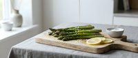 Fresh green asparagus spears arranged on wooden cutting board with lemon slices in bright modern kitchen setting for healthy cooking and meal preparation.